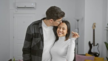 Gülümseyen bir kadın anahtarlarını tutarken, bir erkek yanağından öpüyor, modern bir oturma odasında birlikte yeni bir evi simgeliyor..