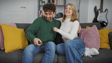 Neşeli bir çift, kadın ve erkek, şık bir oturma odasında uzaktan kumandalı, kanepeli ve gitarlı.