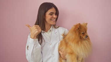 Neşeli genç bir kadın, pomeranian 'ı pembe arka planda tutarken yanlamasına işaret eder.