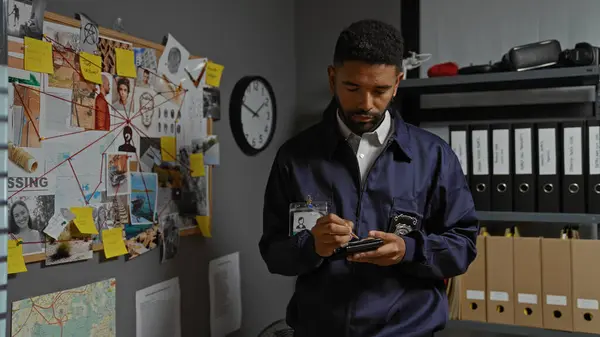 stock image A focused young african american man with a beard taking notes in a detective's office, surrounded by investigation clues and files.