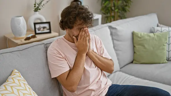 stock image Upset young man sitting on a gray couch at home, covering his face with his hands in a gesture of despair