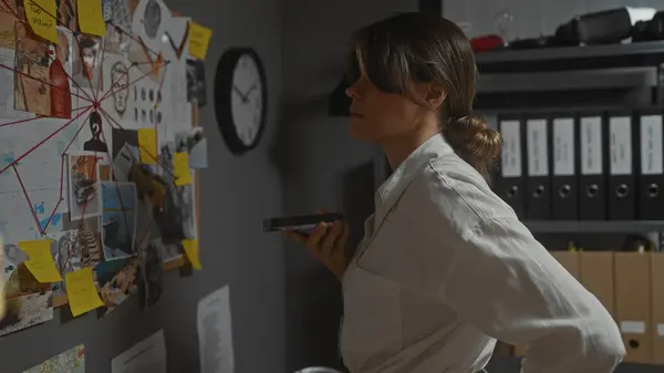 stock image A thoughtful woman detective examines a board of evidence with photos and notes in a dimly lit office.