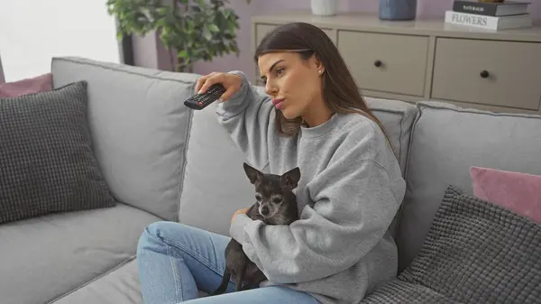 stock image A young woman cuddles with her chihuahua on a sofa at home while holding a remote control.