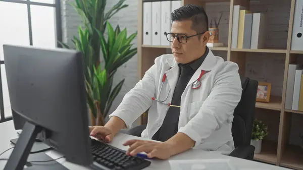 stock image Hispanic male doctor typing on a keyboard in a modern medical office, showcasing healthcare professionals at work.