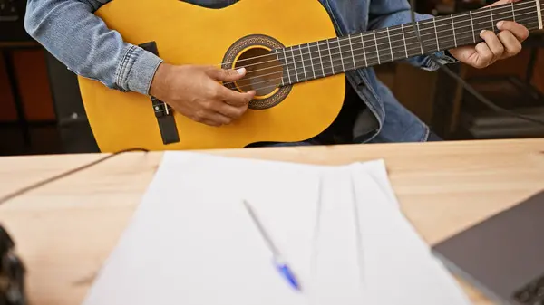 Stock image A close-up of a man's hands playing an acoustic guitar in a music studio with sheet paper and microphone in view.