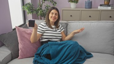 Evdeki kanepede dinlenen genç bir kadın telefon ve battaniyeyle