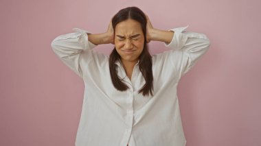 Genç, çekici, İspanyol bir kadın pembe arka planda duruyor. Elleri kulaklarını kapatıyor. Kapalı gözlerle hayal kırıklığını gösteriyor..