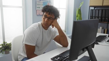 Ofiste oturan genç adam, arka planda organizasyonel materyallerle dolu parlak ve modern bir iş yerinde gözlük takarken bilgisayar ekranına dalgın dalgın bakıyor..