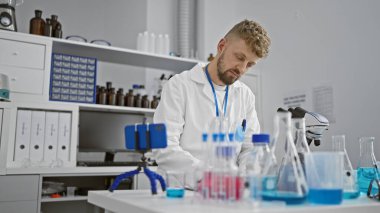 Laboratuvar önlüklü, sakallı, beyaz bir adam modern bir laboratuvar ortamında kimyasalları inceliyor. Araştırmaları temsil ediyor..