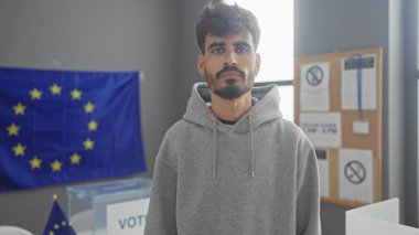 Sakallı bir İspanyol genç adam Avrupa 'da bir seçim merkezinde duruyor ve arkasında bir eu bayrağı var..
