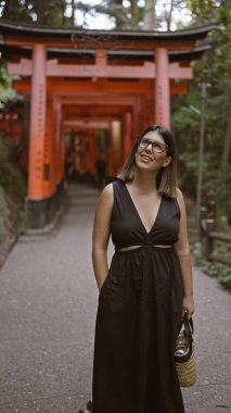Gözlüklü kendine güvenen İspanyol güzeli, kulaktan kulağa sırıtan, Kyoto 'daki Fushimi tapınağındaki turuncu kırmızı torii tüneline huşu içinde bakan.!