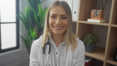 A beautiful young caucasian woman doctor with blonde hair and a stethoscope smiling warmly indoors in a modern clinic or hospital room. clipart