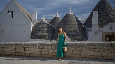 Yeşil elbiseli güzel bir İspanyol kadın İtalya 'nın eski Alberobello kasabasında bir caddede duruyor, arkasında ikonik Trulli evleri olan parlak mavi bir gökyüzünün altında..