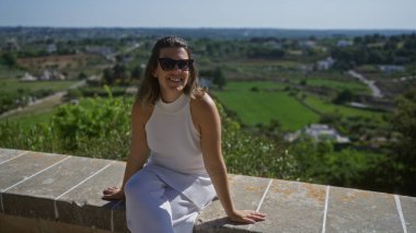 Genç bir İspanyol kadın güneşli bir günde taş bir duvarda otururken Puglia, İtalya 'daki Locorotondo' dan güzel kırsal manzaranın tadını çıkarıyor..