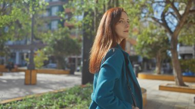 Deniz mavisi ceketli, düşünceli genç bir kızıl saçlı kadın güneşli bir şehir parkı ortamında duruyor..