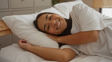Sıcak bir yatak odasındaki yatakta uzanmış gülümseyen bir kadın, rahatlatıcı bir atmosfer sergiliyor..