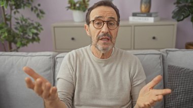 Olgun bir İspanyol adam iyi dekore edilmiş bir oturma odasında kanepede otururken el kol hareketi yapar..