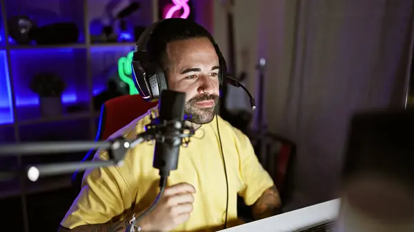 stock image Handsome hispanic man with beard in a gaming room at night, wearing headphones and speaking into a microphone.