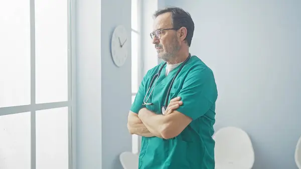 stock image Hispanic male doctor in scrubs with arms crossed standing contemplatively in a well-lit hospital corridor