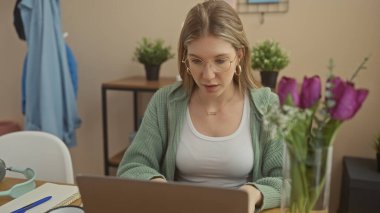 Sarı saçlı ve gözlüklü, içi mor lalelerle süslenmiş rahat bir evde dizüstü bilgisayarda çalışan odaklanmış genç bir kadın.