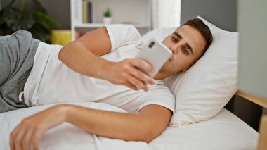 Genç İspanyol adam yatak odasında akıllı telefon kullanıyor. Evde rahat ve rahat görünüyor..