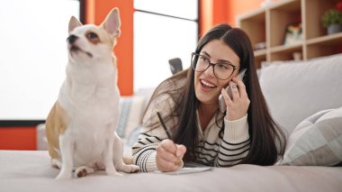 Chihuahua köpekli genç İspanyol kadın telefonda konuşuyor ve evdeki kanepede uzanmış notlar alıyor.