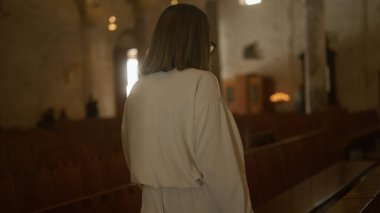 Genç bir İspanyol kadın İtalya 'da bir kilisede duruyor, sırtı kameraya dönük, açık renkli bir bluz ve gözlük takıyor, arka planda loş ışıklandırma ve dini dekor var..