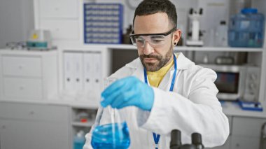 Sakallı İspanyol adam laboratuarda güvenlik gözlüğü ve eldiven takarak deney yapıyor..