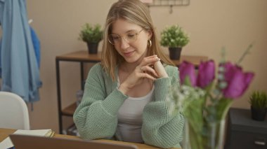 Gözlüklü, gülümseyen bir kadın evinde oturmuş, elinde çiçekler ve laptopuyla rahat bir atmosferi çağrıştırıyor..