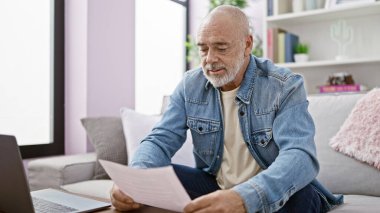 Gri saçlı, sakallı, kot ceket giyen son sınıf öğrencisi evde olgunluk ve ev hayatını gösteren bir belge okuyor..