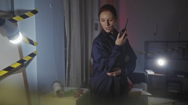 Odaklanmış kızıl saçlı bir polis memuru loş ışıkta yanan bir suç mahallini araştırıyor ve telsizle yardım istiyor..