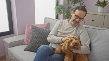 Orta yaşlı bir adam oturma odasındaki kanepede dinlenirken kaniş köpeğini sevgiyle okşar..