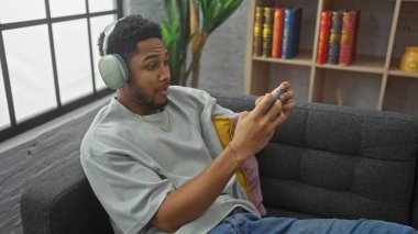 Afrika kökenli Amerikalı bir adam oturma odasında video oyunları oynuyor, kulaklık takıyor, rahat, rahatlatıcı, teknoloji, eğlence, konforlu oyun, kanepe ve iç mekan..