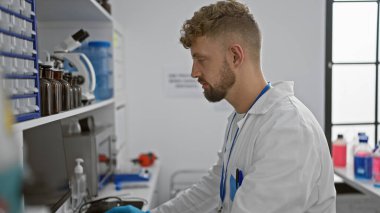 Sakallı beyaz bir adam tıbbi ekipman ve şişelerle çevrili bir laboratuvar ortamında özenle çalışıyor..