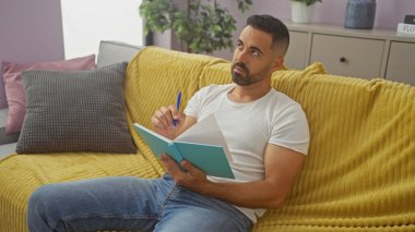 Düşünceli İspanyol bir adam sarı bir kanepeye oturur, rahat bir oturma odasında, bir deftere yazarak yaratıcılık ve rahatlık uyandırır..