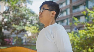 Düşünceli, beyaz gömlekli genç bir Asyalı sakin bir şehir ortamında yeşilliğin ortasında dikiliyor..