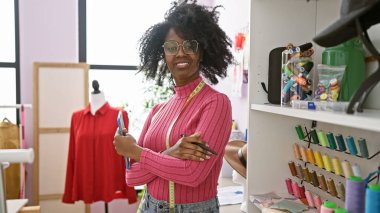 Kolları çapraz bağlanmış, kendine güvenen Afrikalı bir kadın. Renkli ipliklerle çevrili bir terzi dükkanında duruyor. Bir de manken..