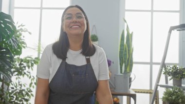 Çiçekçide gülümseyen orta yaşlı İspanyol bir kadın. Etrafı bitki ve yeşilliklerle çevrili bir önlük giyiyor..