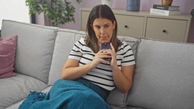 Güzel, esmer, genç, İspanyol bir kadın oturma odasındaki koltukta oturmuş, elinde cep telefonu tutuyordu..