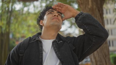 Kıvırcık saçları olan genç İspanyol bir adam güneşli bir park manzarasına bakıyor, düşünceli bir şehir kaşifi havası veriyor..