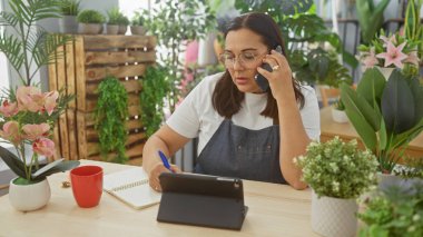 İspanyol bir kadın çiçekçi dükkanında yeşil bitkiler ve çiçeklerle çevrili bir defterde yazarken bir telefon görüşmesi yapıyor..