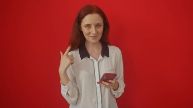 Beyaz bluzlu kızıl saçlı kadın kırmızı arka planda akıllı telefonu tutarken yukarı bakıyor.