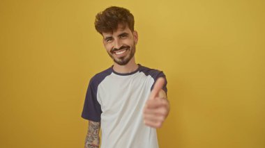 Sakallı, gülümseyen İspanyol adam sarı arka planda izole edilmiş baş parmağını kaldırıyor.