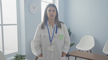 Stetoskop ve rozetle klinik odasında duran kendine güvenen genç kadın doktor, profesyonelliği resmediyor..