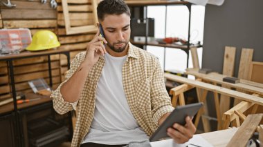 Sakallı bir adam marangozluk atölyesinde bir tablet tutarken telefonla konuşuyor..