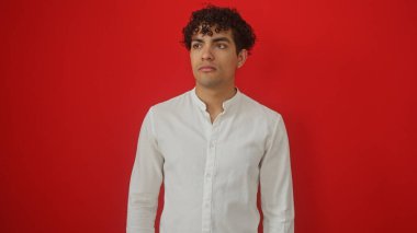 Kıvırcık saçlı, beyaz tişörtlü, parlak kırmızı arka planlı, düşünceli ve kendine güvenen genç İspanyol bir adam..