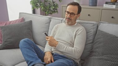 Yakışıklı, orta yaşlı İspanyol bir adam rahat bir oturma odasında, elinde uzaktan kumandayla kanepede dinleniyor, gözlük takıyor ve rahat giyiniyor, yumuşak yastıklarla çevrili..