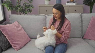 Latin bir kadın, rahat bir oturma odasında, kanepede beyaz bir köpekle oynuyor.
