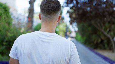 Beyaz tişörtlü genç bir İspanyol adam, bulanık bir sokak geçmişi olan yemyeşil bir parkta düşünceli bir şekilde dikiliyor..