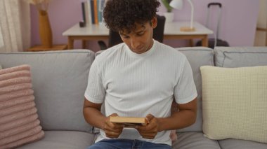 Beyaz tişörtlü ve modern dekorlu, rahat bir oturma odasında kanepede oturmuş kitap okuyan genç İspanyol bir adam..